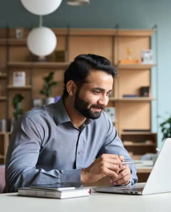 A bearded man in a shirt sits at a desk, working on a laptop.