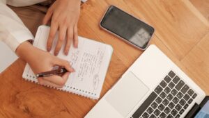 A person writing notes in a notebook with a pen, while a smartphone and a laptop are placed on a wooden desk, symbolizing email writing or professional communication.